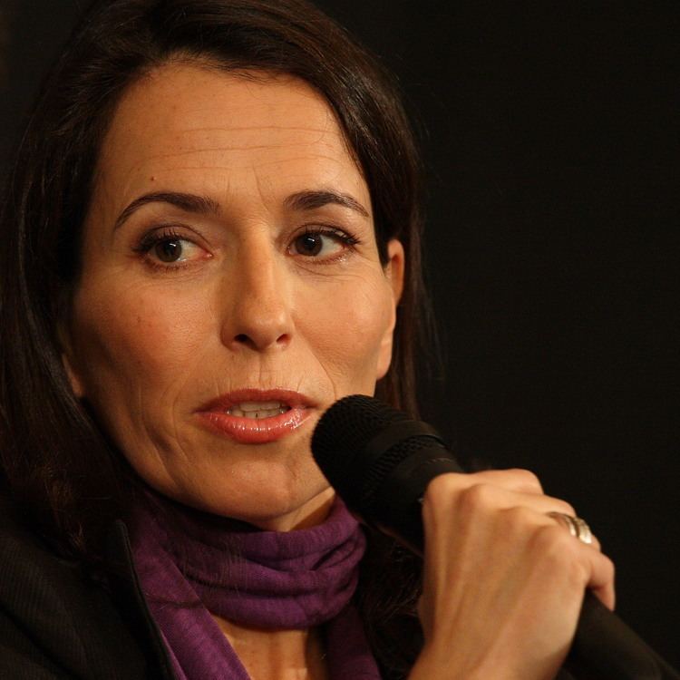 Anne Will speaking while holding the microphone and wearing a violet scarf, black blouse, and ring
