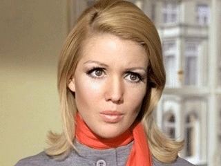 Annette Andre looking at something while wearing an orange scarf and gray blouse