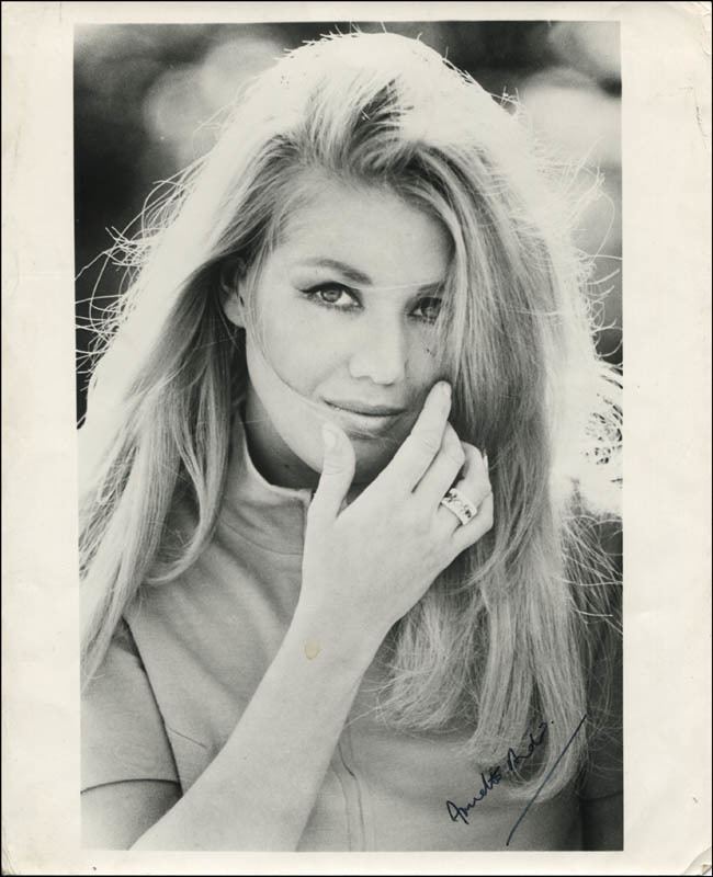 Annette Andre with a tight-lipped smile and hand on her face while wearing a blouse and ring and an autograph on the lower right side