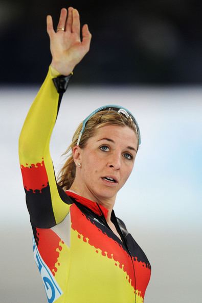 Anni Friesinger-Postma with a serious face while raising her hand, with blonde hair, a protective eyeglass on her head, wearing earrings, rings, and a multi-colored bodysuit.