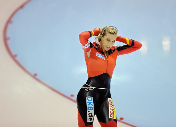Anni Friesinger-Postma with a serious face while tying her blonde hair, with a protective eyeglass on her head, and wearing a multi-colored bodysuit with logos.