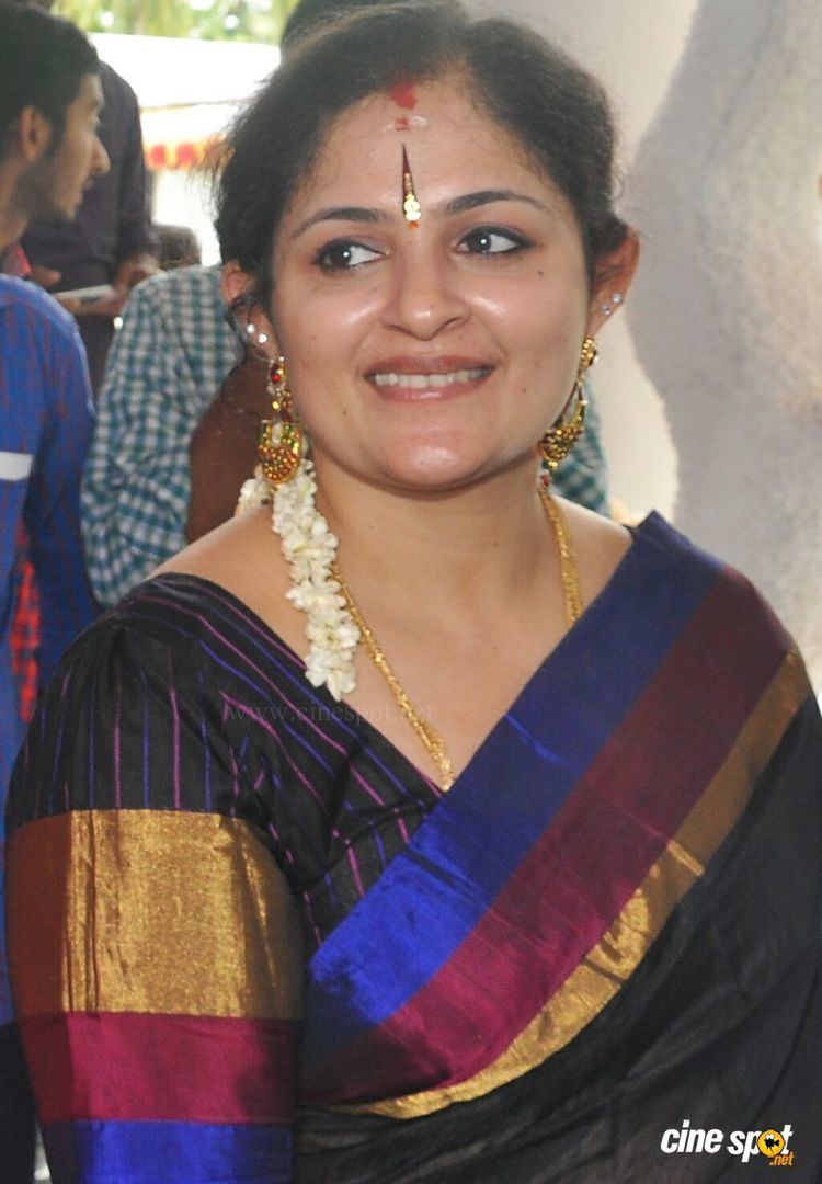 Annie smiling with bindis on her forehead while wearing gajra, earrings, necklace, black dress with a combination of the color blue, gold, and maroon
