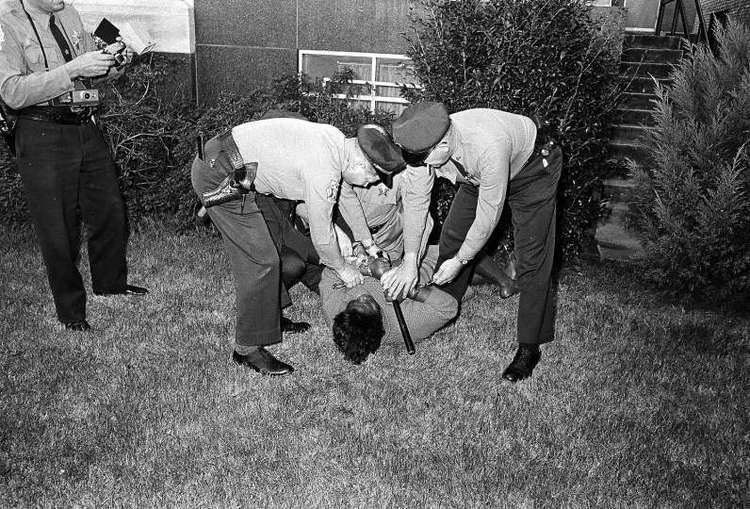 Oprah Winfrey as Annie Lee Cooper lying and resisting on the grassy ground while holding a billy club while being forcefully arrested and handcuffed by three uniformed policemen with plants and a window in the background. Oprah is wearing a long-sleeve dress while the three policemen are wearing long-sleeve uniforms, slacks, belts with guns in their holsters, socks, and formal shoes. On the left is a uniformed policeman holding a portable flash and a camera hanging on his neck in a movie scene from "Selma", a 2014 American historical drama film. He has a belt with a gun in its holster and is wearing a long-sleeved uniform with a necktie and a pair of slacks.