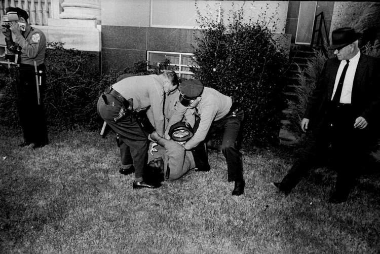 Oprah Winfrey as Annie Lee Cooper lying and resisting on the grassy ground while holding a billy club while being forcefully arrested and handcuffed by three uniformed policemen with plants and a window in the background in a movie scene from "Selma", a 2014 American historical drama film. Oprah is wearing a long-sleeve dress while the three policemen are wearing long-sleeve uniforms, slacks, belts with guns in their holsters, socks, and formal shoes. On the left is a uniformed policeman wearing a long-sleeved uniform with a necktie, a pair of slacks, a belt with a gun in its holster; holding a portable flash and a camera hanging on his neck. On the right is a man has a cap paired and black shoes, wearing a black tuxedo over a white collared shirt and a black necktie.