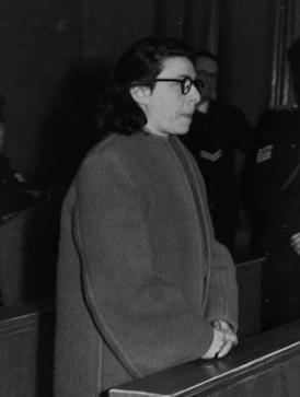 Ans van Dijk standing while moving her lips and holding her hand with a man standing in his background, he has short black hair wearing black eyeglass and a long zip coat.