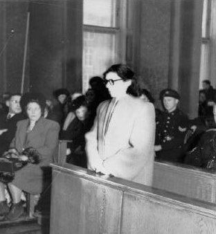 Ans van Dijk standing in front while holding her hand around people sitting in his background including policemen. Ans has short hair wearing black eyeglass and a long coat.