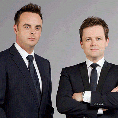In a gray background, from left, Anthony McPartlin is serious, standing, hands down has black hair, black eyebrows wearing a white polo with black necktie under a black coat at the right, Declan Donnelly is serious, standing, arms crossed has brown hair wearing a silver watch, a white polo with black necktie under a black coat.
