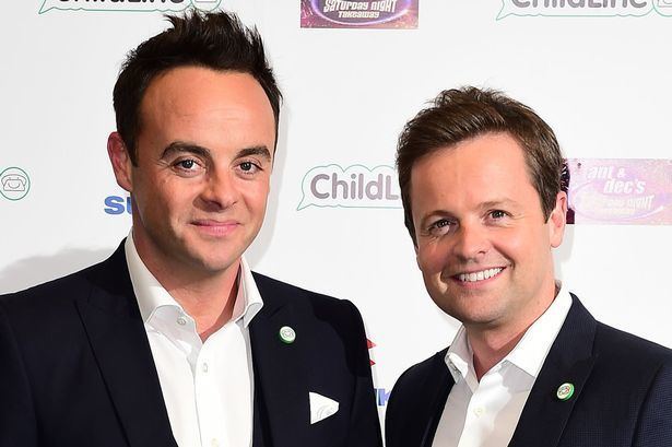 In a white printed backdrop, from left, Anthony McPartlin is smiling, standing, has black hair, black eyebrows wearing a white polo under a black coat with a printed pin on his left chest with white scarf, at the right, Declan Donnelly is smiling, standing, has brown hair wearing a white polo under a black coat with a printed white pin on his left chest.