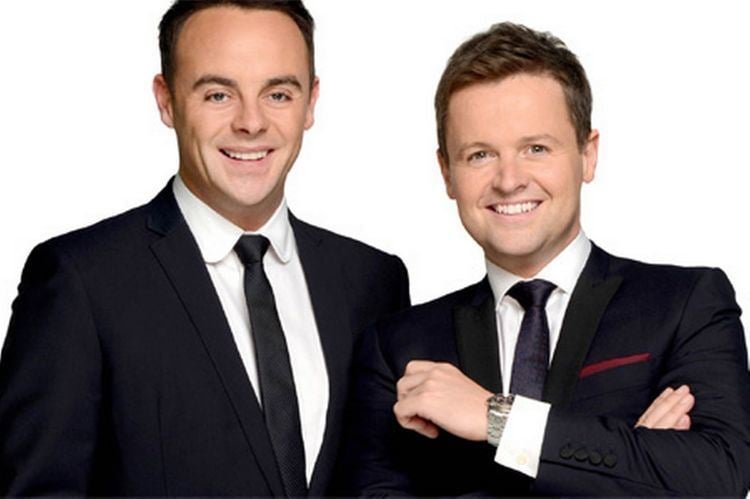 In a white background, from left, Anthony McPartlin is smiling, standing, hands down has black hair, black eyebrows wearing a white polo with a black necktie under a black coat at the right, Declan Donnelly is laughing, standing, leaning to his right, arms crossed has brown hair wearing a silver watch, a white polo with black necktie under a black coat.