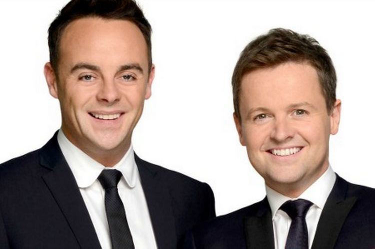 In a white background, from left, Anthony McPartlin is smiling, standing has black hair, black eyebrows wearing a white polo with a black necktie under a black coat at the right, Declan Donnelly is laughing, standing, leaning to his right, has brown hair wearing a white polo with black necktie under a black coat.