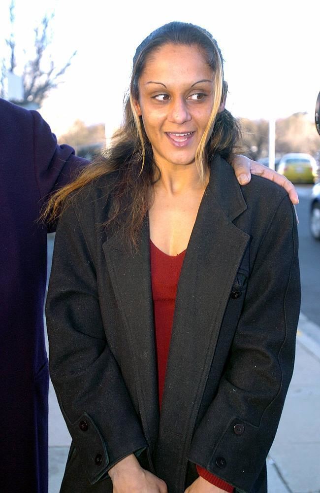 Anu Singh smiling mischievously while looking on her left and a man beside her is holding her shoulder and cars and a bare tree in the background. She has a half up-do hairstyle with blonde highlights, a drawn eyebrows, and wearing a red knitted-long sleeve blouse under a black coat with a visible cleavage.