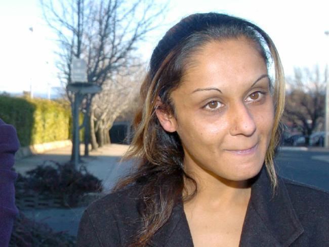 Anu Singh biting her lower lip while there are bare trees and a lamp post in the background, leaving the ACT Supreme Court in 2004. Anu Singh with a half up-do hairstyle and blonde highlights, drawn eyebrows, wearing a black coat.
