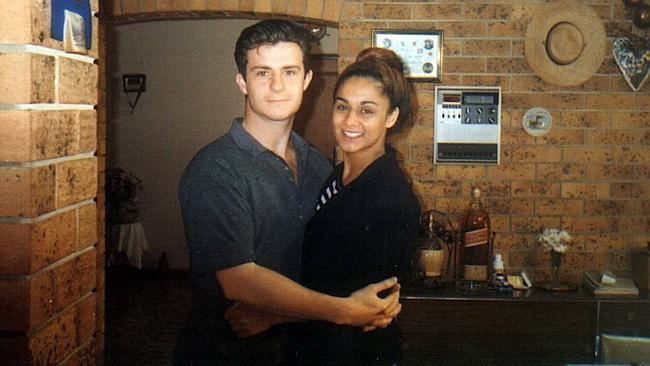A photo of Anu Singh with her boyfriend, both are smiling while hugging each other with a door, a brick wall, picture frames, a hat, a circuit breaker on the wall and a table-drawer with a Red Label bottle and a flower vase in the background. Anu Singh in a bun hair is wearing a black and white striped blouse under a black blazer while Joe is wearing a dark gray collared-polo shirt.