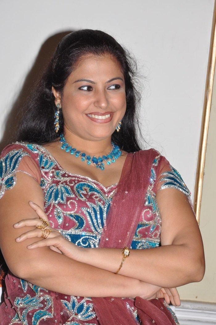 Anusha smiling while wearing a maroon and blue dress, blue necklace, earrings, gold ring, and wristwatch