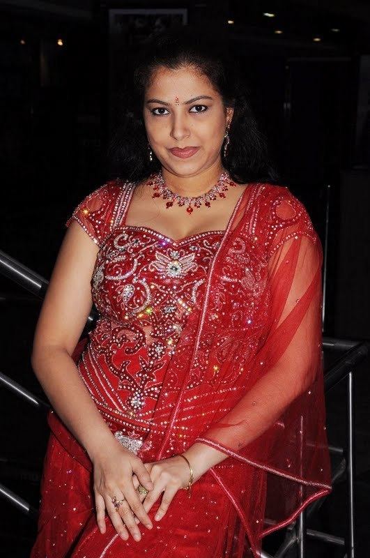 Anusha smiling while wearing a red dress with a sequence, red necklace, earrings, bracelet, and ring