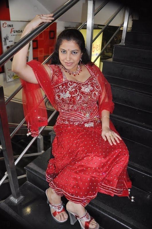 Anusha smiling and sitting on the stairs while wearing a red dress with a sequence, red necklace, and earrings