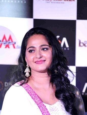 Anushka Shetty with a tight-lipped smile and black curly hair while wearing a white and pink dress and earrings