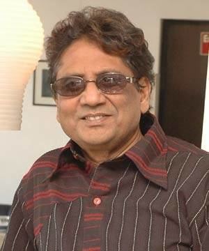 Anwar (singer) smiles while wearing a brown collared long-sleeved shirt and pair of eyeglasses inside a room