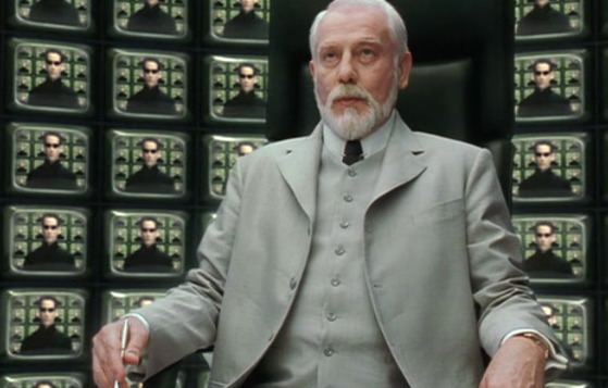 Helmut Bakaitis sitting on a chair and wearing a gray vest, gray coat, white sleeves in a scene from the film "The Architect Matrix" in 2003