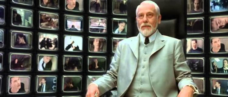 Helmut Bakaitis sitting on a chair and wearing a gray vest, gray coat, white long sleeves, and black necktie in the film "The Architect Matrix" in 2003