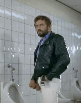 Armin Meier (actor) is serious, has brown hair, a beard, and a mustache, standing inside the comfort room in front of a urinal, right hand holding his penis, left hand on his pocket, wearing a black belt, a blue shirt under a black jacket and white pants.