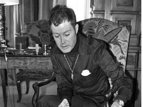 Arndt von Bohlen und Halbach sitting inside a room wearing a formal suit, a necklace and a watch.