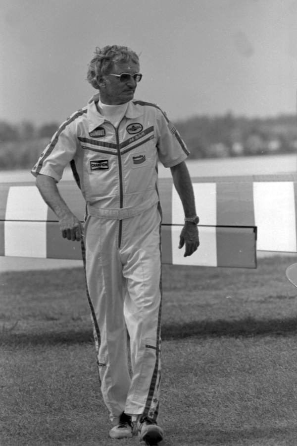 Stunt pilot Arthur Everett Scholl at the Homestead General Aviation Airport during an air show, has white hair, wearing black shades, a wristwatch on his left hand, and his uniform a white polo with black strips on the shoulder and chest with his name, white pants with black strips on the side, and white shoes.