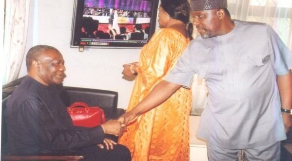 Arthur Nzeribe smiling while sitting on a couch and doing a handshake with a man standing in front of him with a woman at the back. Arthur is wearing a black jalabiya while the man is wearing a gray kufi and gray juba thobe while the woman at the back is wearing an orange saree dress.
