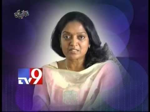 In TV9 with a blue background, Aruna Mucherla is serious, has black long hair, a bindi on forehead, wearing a gold necklace, earrings and white salwar kurta.