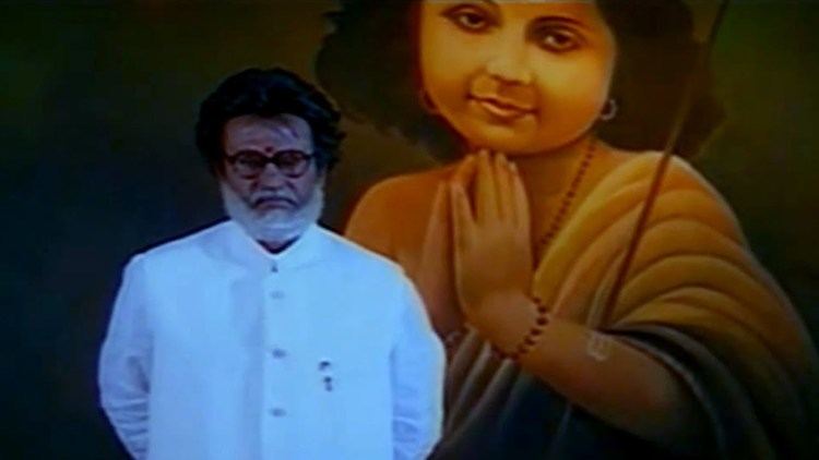 Rajinikanth with a serious face, standing in front of a photo of Lord Murugan with beard and mustache, wearing eyeglasses, and a white stand collar jacket.