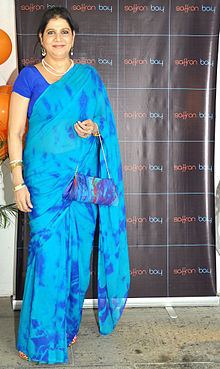 Asha Sachdev smiling while holding a blue bag and wearing a blue dress and pieces of jewelry