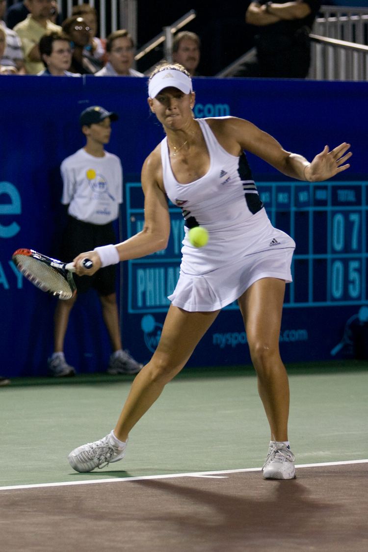 Ashley Harkleroad is serious while hitting the tennis ball with her right hand, with the audience behind her, she is wearing earrings, a necklace, and a sweatband on her right hand, a  bracelet on her left-hand black and white Adidas tank top, a white Adidas skort, a white adjustable Adidas visor cap, white socks, and shoes. Behind her is a ball boy, wearing a white printed racket shirt, black shorts, a black cap, white socks, and gray-black shoes.
