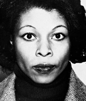 Assata Shakur with a serious face, short hair, and wearing a gray coat over a black shirt.