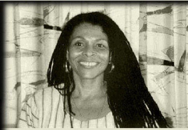 Assata Shakur smiling, with long dreadlocks hair, wearing loop earrings, a necklace, and a white striped top.