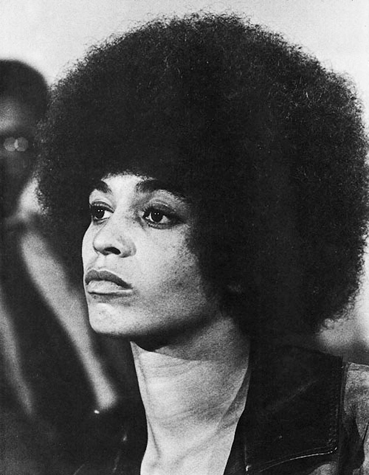 Angela Davis with a serious face while looking at something, with kinky hair and wearing a black blouse.