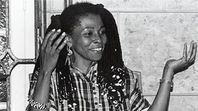 Assata Shakur is smiling while moving her hands, with dreadlocks hair, wearing earrings, a bracelet, a watch, and a checkered blouse.