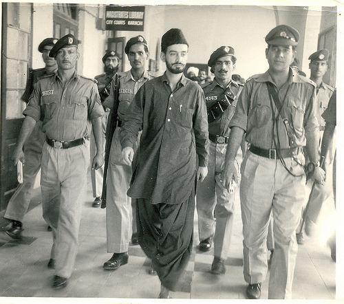 Sardar Ataullah Mengal walking with the policemen while wearing a cap, long sleeves, and pants