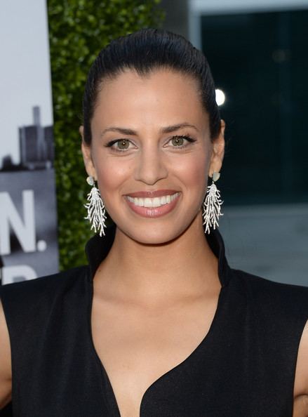 Athena Karkanis smiling with tied-up hair while wearing earrings and a black sleeveless blouse with a plunging neckline