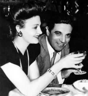 Athina Livanos smiling while holding a drink and wearing a black dress, necklace and earrings togther with Alexander Onassis