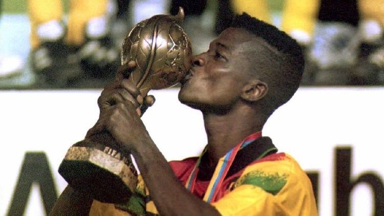 Awudu Issaka is proud, holding up a copper trophy with his hands, kissing it with his lips, standing sideways right shoulder at the back, wearing a medal with yellow,blue and red colored lace, a yellow with green and orange printed polo shirt.