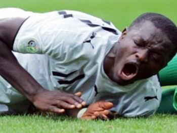 Awudu Issaka is in pain, lying on the ground of the football field, mouth half open, eyes closed, wearing a white with black line football uniform with a printed silhouette of a Jaguar on shoulders. At the back is a leg wearing a green socks.