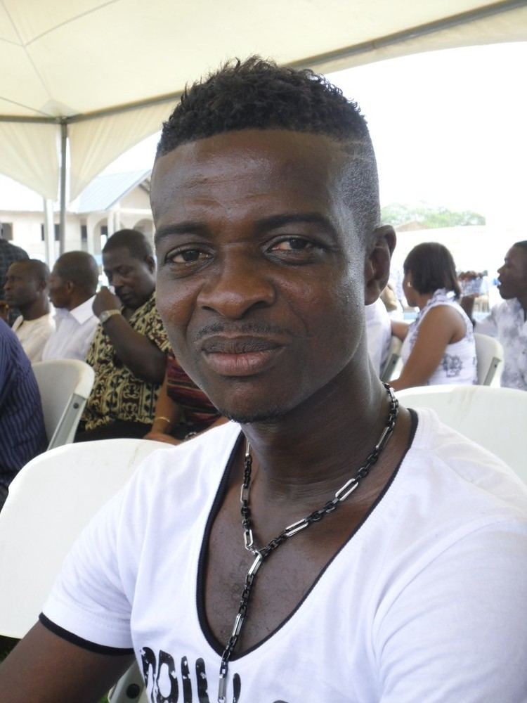 Awudu Issaka is serious, sitting in a white chair, has short black hair, wearing a black bead with silver necklace and a white shirt, at the back is group people sitting on white chair under a white tent, from left a man is serious, has bald hair wearing a white polo, 2nd from left a man, is serious looking at his right, has bald hair, wearing a white polo, 3rd from left a man is serious, sitting looking down, eyes closed right hand closed to his face, has bald hair wearing a gold watch and a yellow printed polo shirt, 4th from left, a woman, sitting on a white chair, looking at the back has short black hair wearing a gold earings and a white sleeveless top, at the right a man is serious, sitting, mouth half open, looking at his right, has bald hair wearing a white printed polo shirt.
