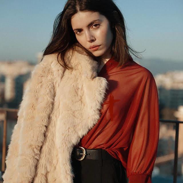 Aya Wolf with a serious face, with short hair, wearing a cream fur blazer, orange long sleeve blouse, black belt, and black pants.