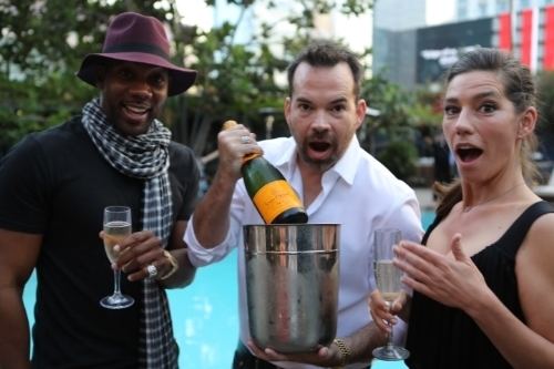 Ayisha Davies holding a glass of wine and wearing a black top while the two men beside her holding a wine