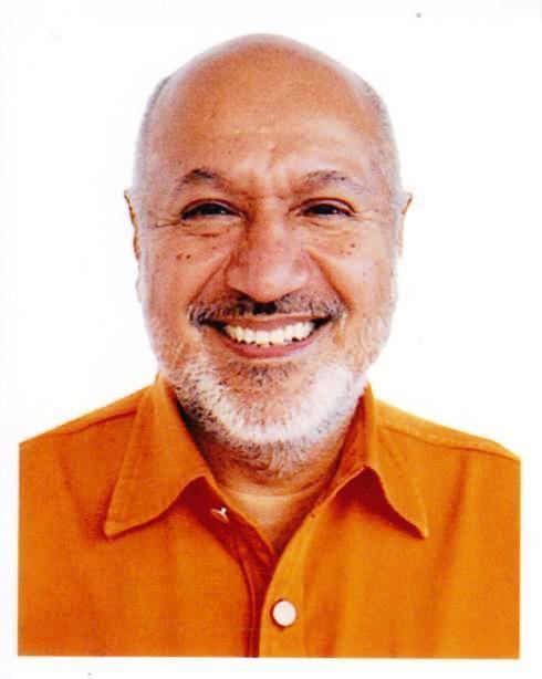 On a white background, Aziz Mohammad Bhai is smiling, has a bald head, a white beard and a mustache, wearing an orange polo with white buttons.