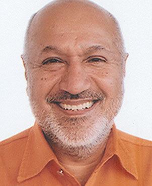 On a white background, Aziz Mohammad Bhai is smiling, has a bald head, a white beard and a mustache, wearing an orange polo.