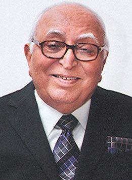 On a white background, Aziz Mohammad Bhai is smiling, has a bald top head and a white side hair a white eyebrows wearing eyeglasses, white polo with black checkered necktie under a black suit with checkered handkerchief.
