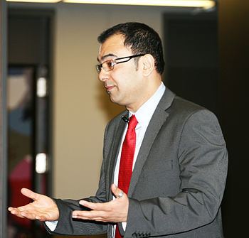 Azmi Haq with a serious face, wearing eyeglasses, gray suit, white long sleeves, and a red tie.