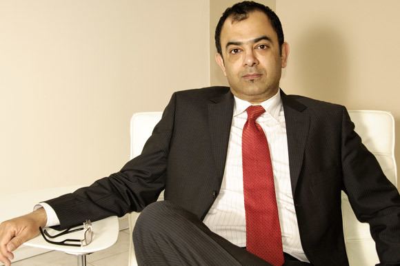 Azmi Haq with a serious face while sitting on a white couch, his right hand is on the small table with his eyeglasses, wearing a black suit, white long sleeves, red tie, and black pants.