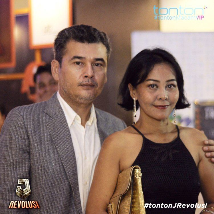 Azri Iskandar with a serious face while holding her wife’s left shoulder, Ellie Suriaty. Azri has black hair, a beard, and mustache wearing a white polo under a gray coat and Ellie is smiling with a gold purse in her arm, she has black hair wearing a dangle earring and a black sexy top